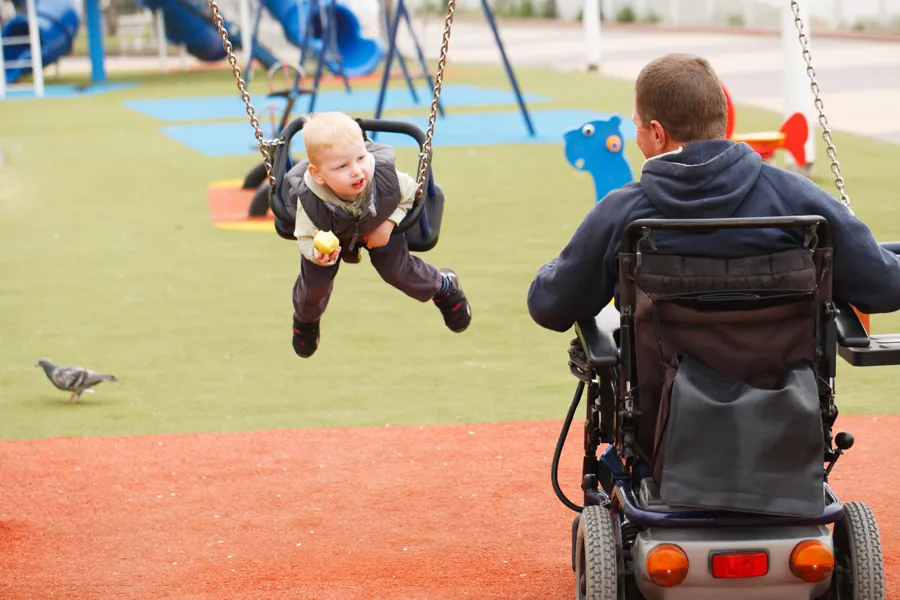 Far som sitter i elektrisk rullestol dytter barn på huske