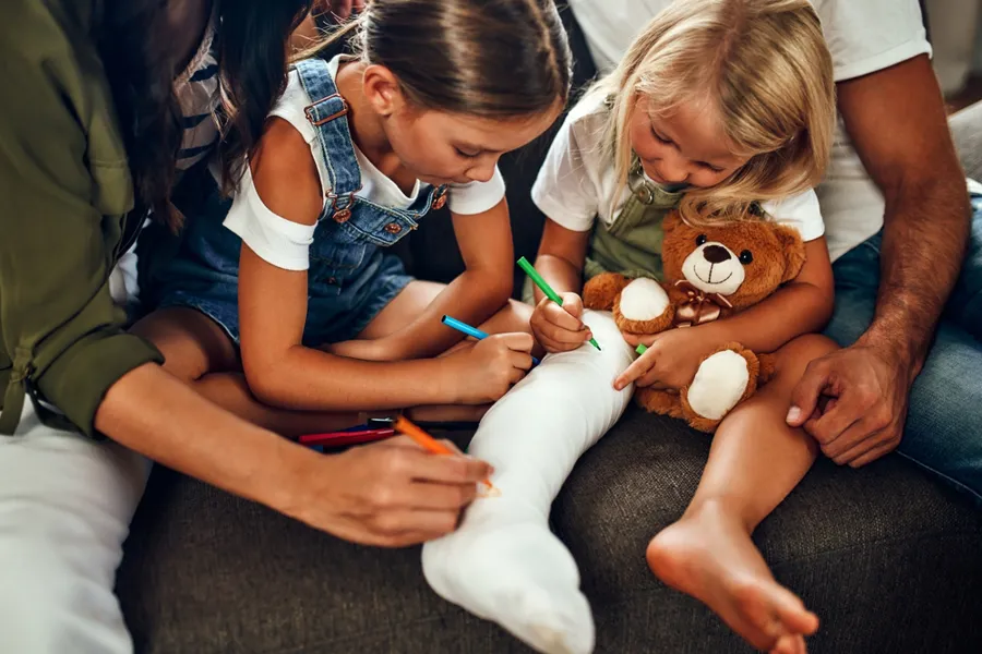 To barn som tegner og leker med en bamse. Det ene barnet har gips på benet