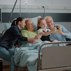 En familie sitter i en sykehusseng, mor, barn og besteforeldre. Barnet tar selfie med en smarttelefon.