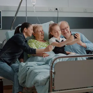 En familie sitter i en sykehusseng, mor, barn og besteforeldre. Barnet tar selfie med en smarttelefon.
