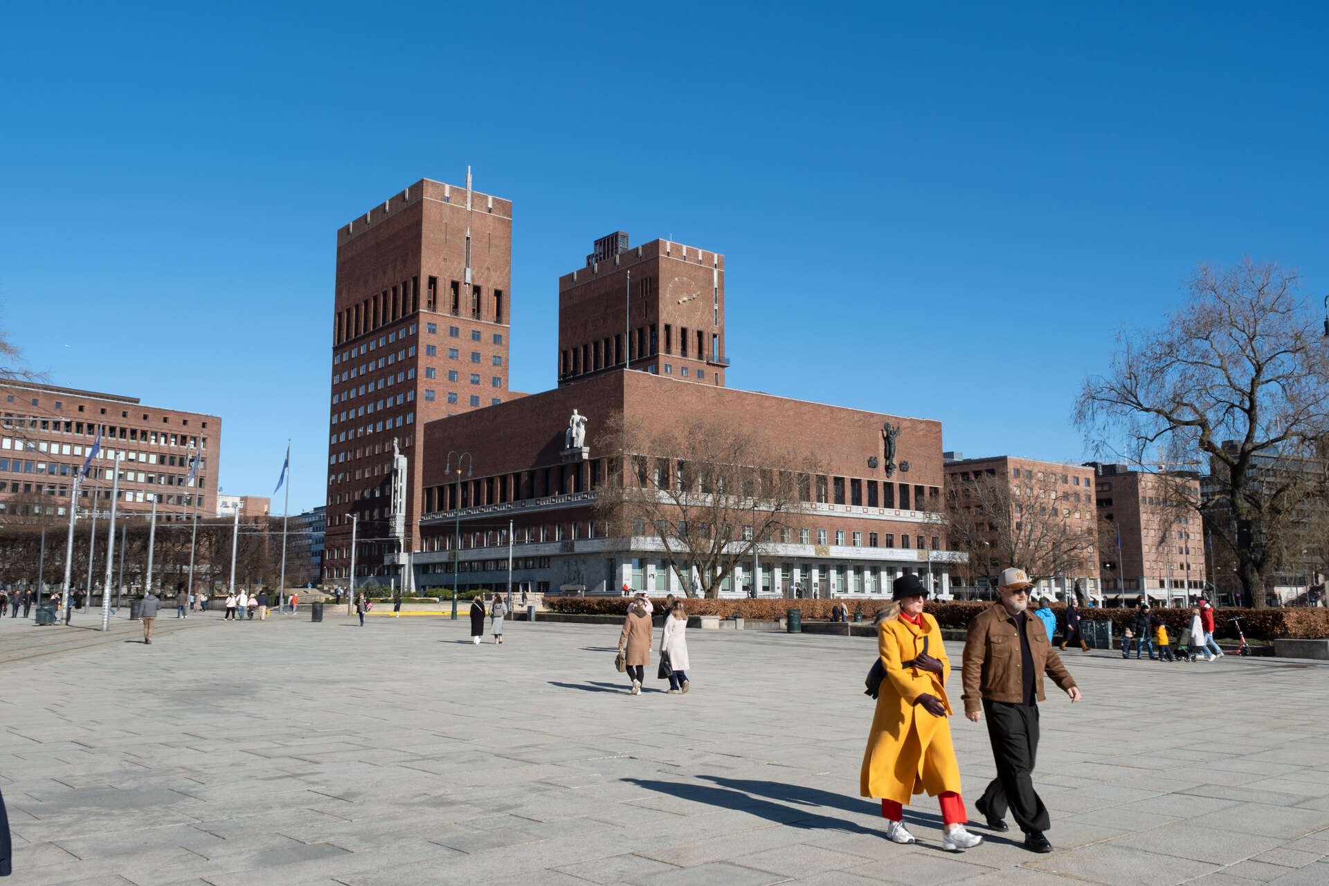 A group of people walking in a courtyard with buildings in the background with Oslo City Hall in the background