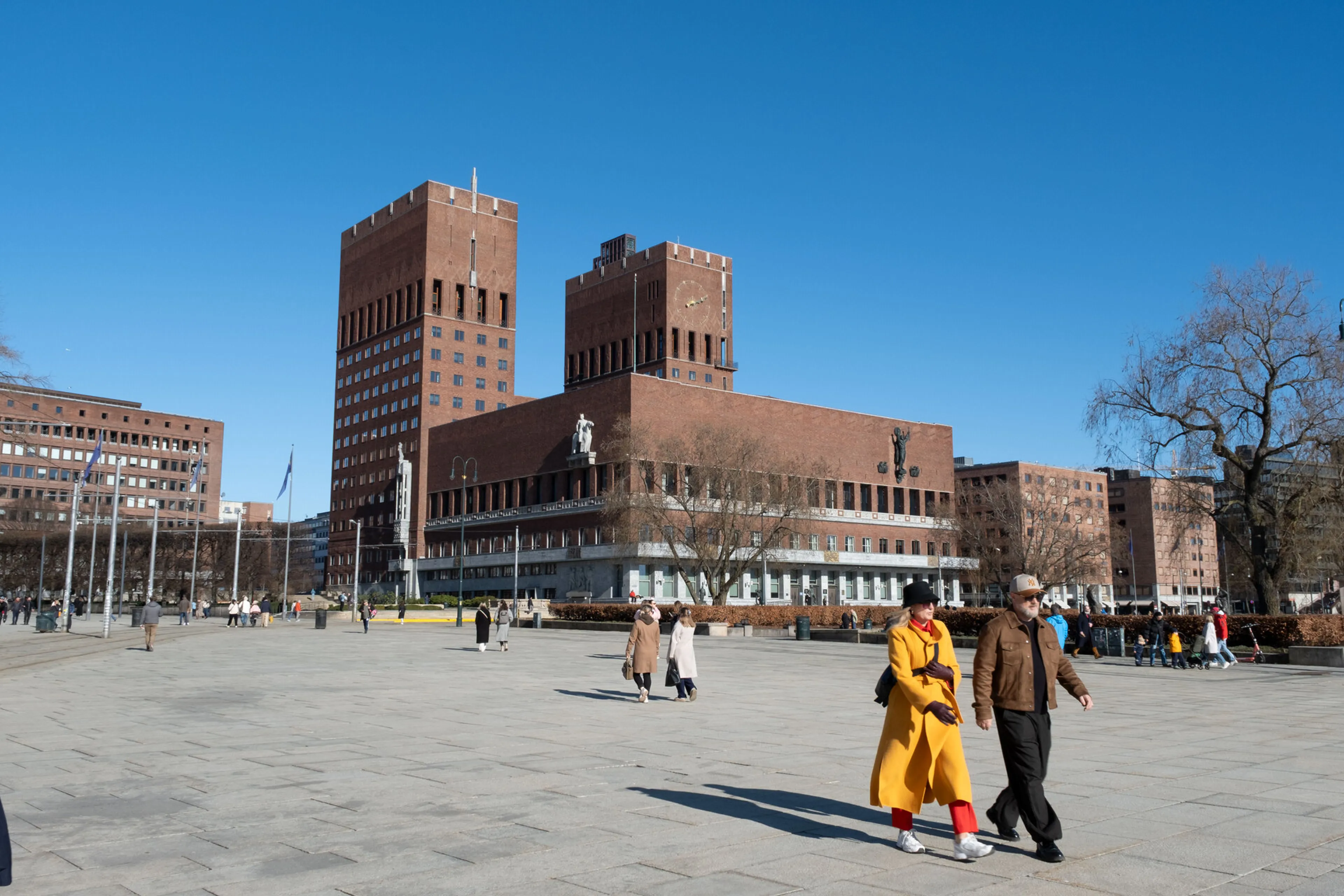 A group of people walking in a courtyard with buildings in the background with Oslo City Hall in the background
