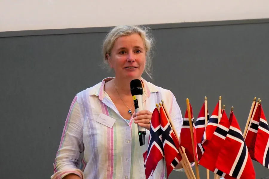 Annette Halvorsen på talerstolen. Hun holder en mikrofon. Det står norske flagg i en vase foran henne