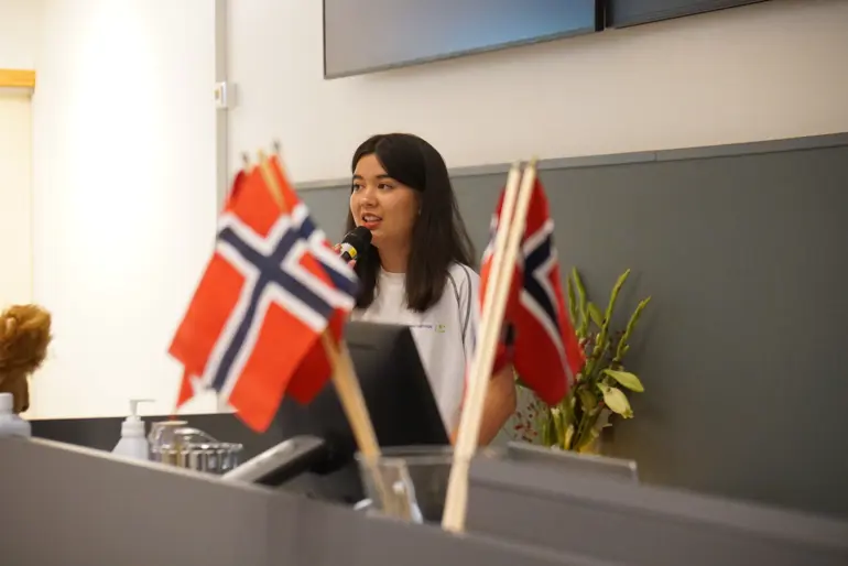 Julie snakker i en mikrofon på talerstolen. Det står norske flagg foran henne