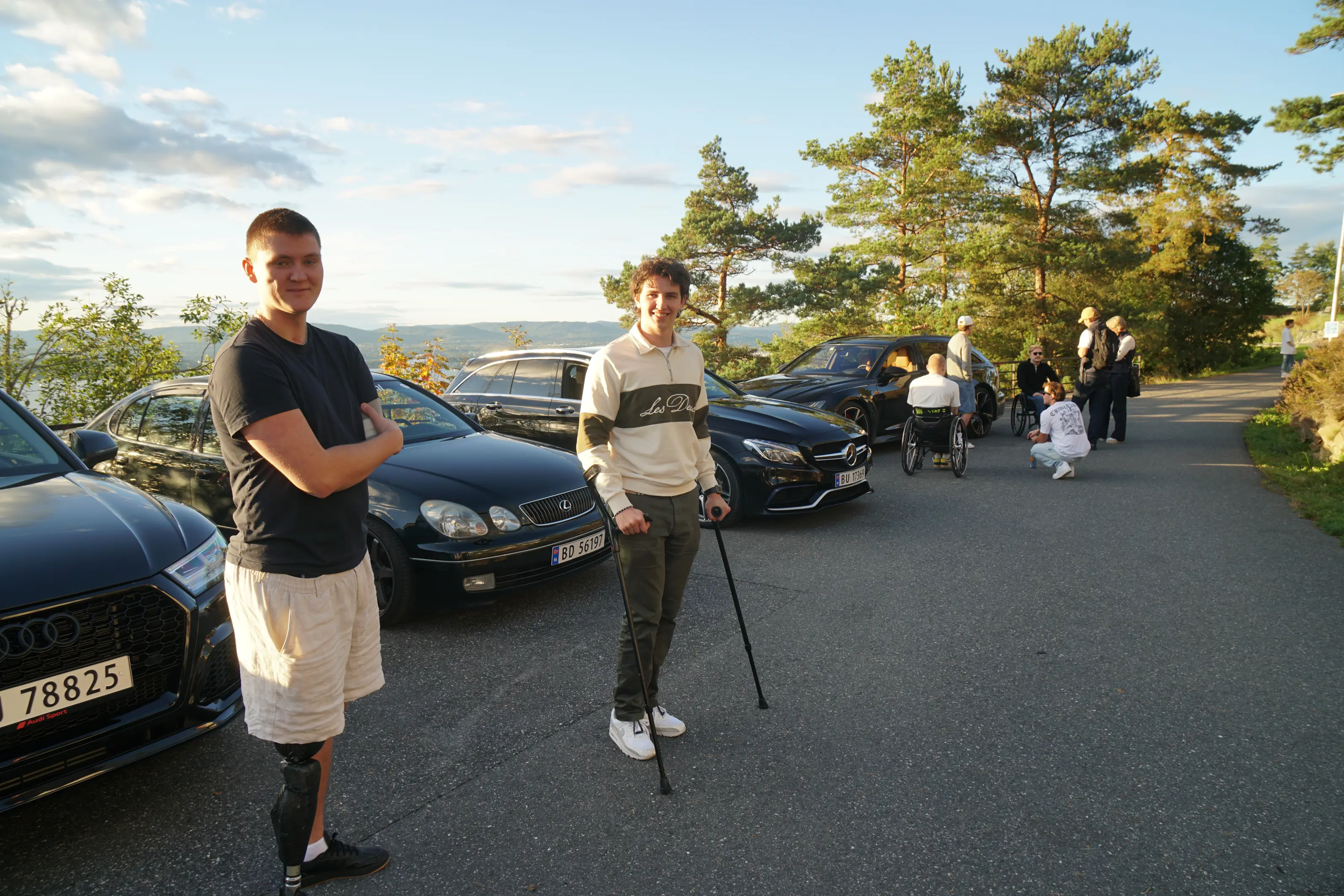 Victor og Jonas står foran noen flotte biler. Begge to har funksjonsnedsettelser. I bakgrunn står en gruppe mennesker hvorav to rullestolbrukere