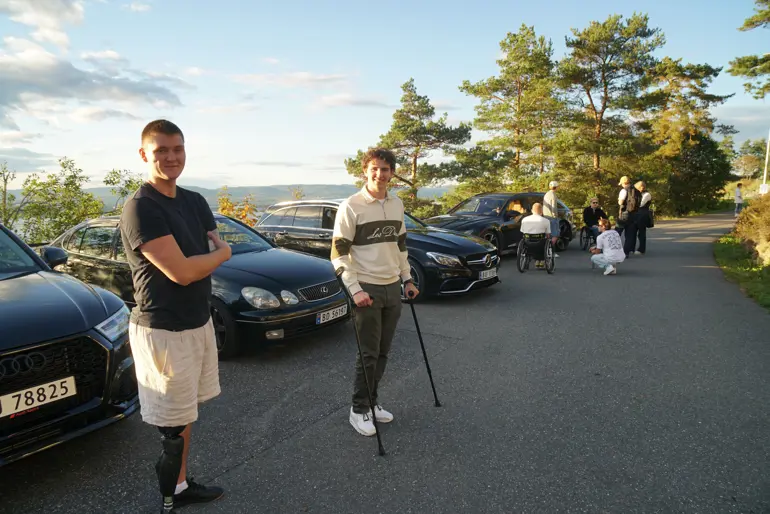 Victor og Jonas står foran noen flotte biler. Begge to har funksjonsnedsettelser. I bakgrunn står en gruppe mennesker hvorav to rullestolbrukere