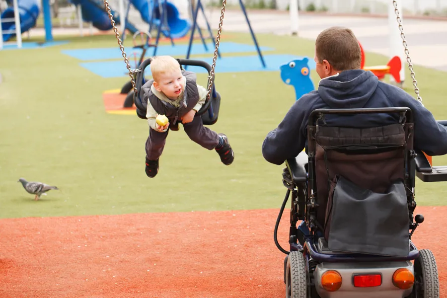 Far som sitter i elektrisk rullestol dytter barn på huske