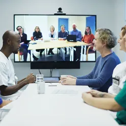 Flem mennesker sitter rundt et møtebord i et møte, de ser på en skjerm hvor det sitter seks andre mennesker som er med på møtet