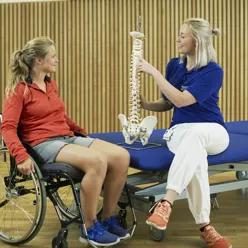En behandler som holder en modell av en ryggrad og viser den til en kvinne i rullestol