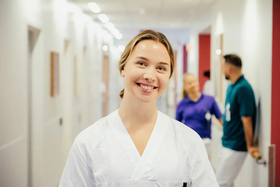 En sykepleier smiler mot kamera, i bakgrunnen står en fysioterapeut og en ergoterapeut.