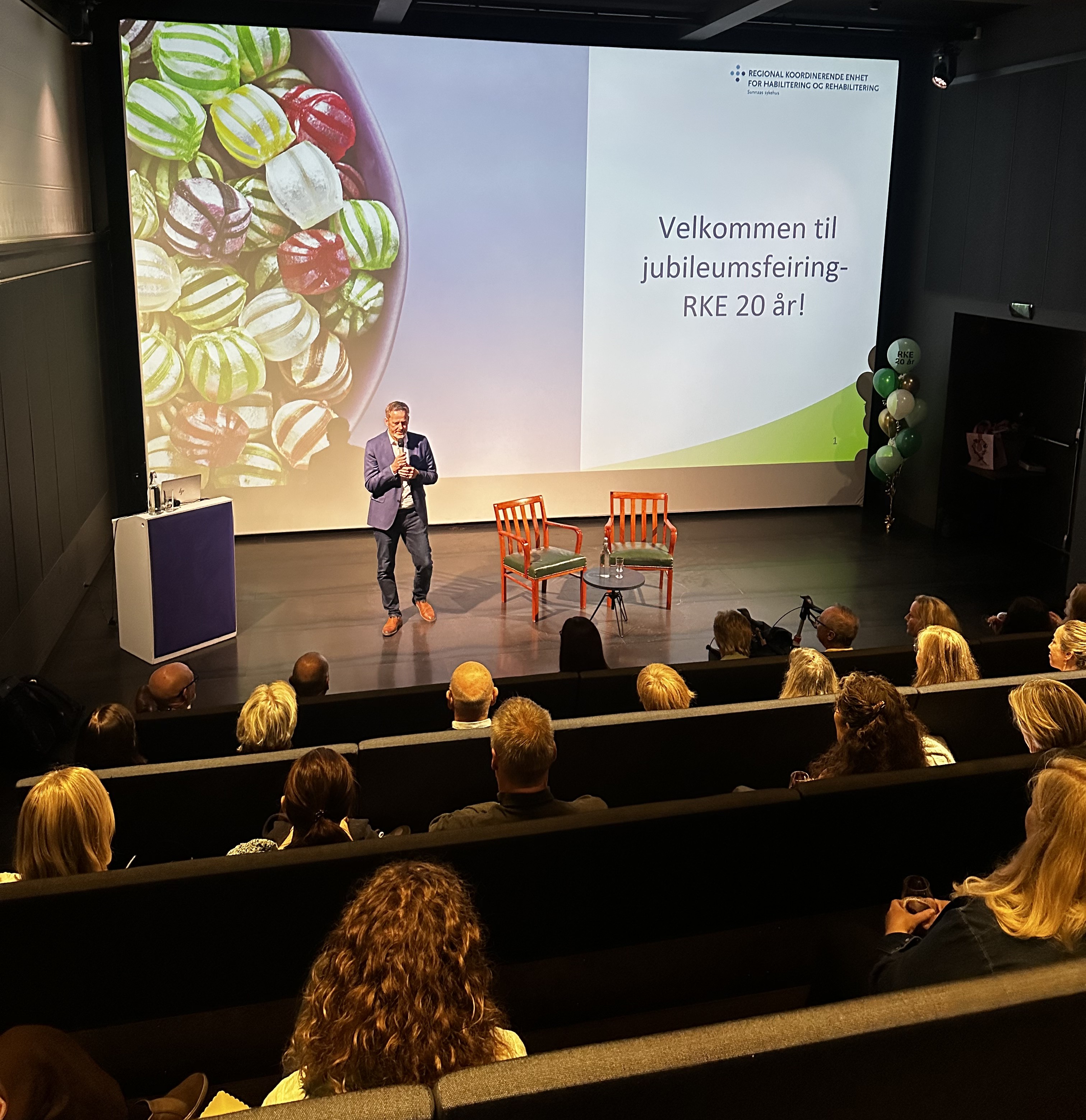 En person som står på en scene med en gruppe mennesker som sitter i stoler