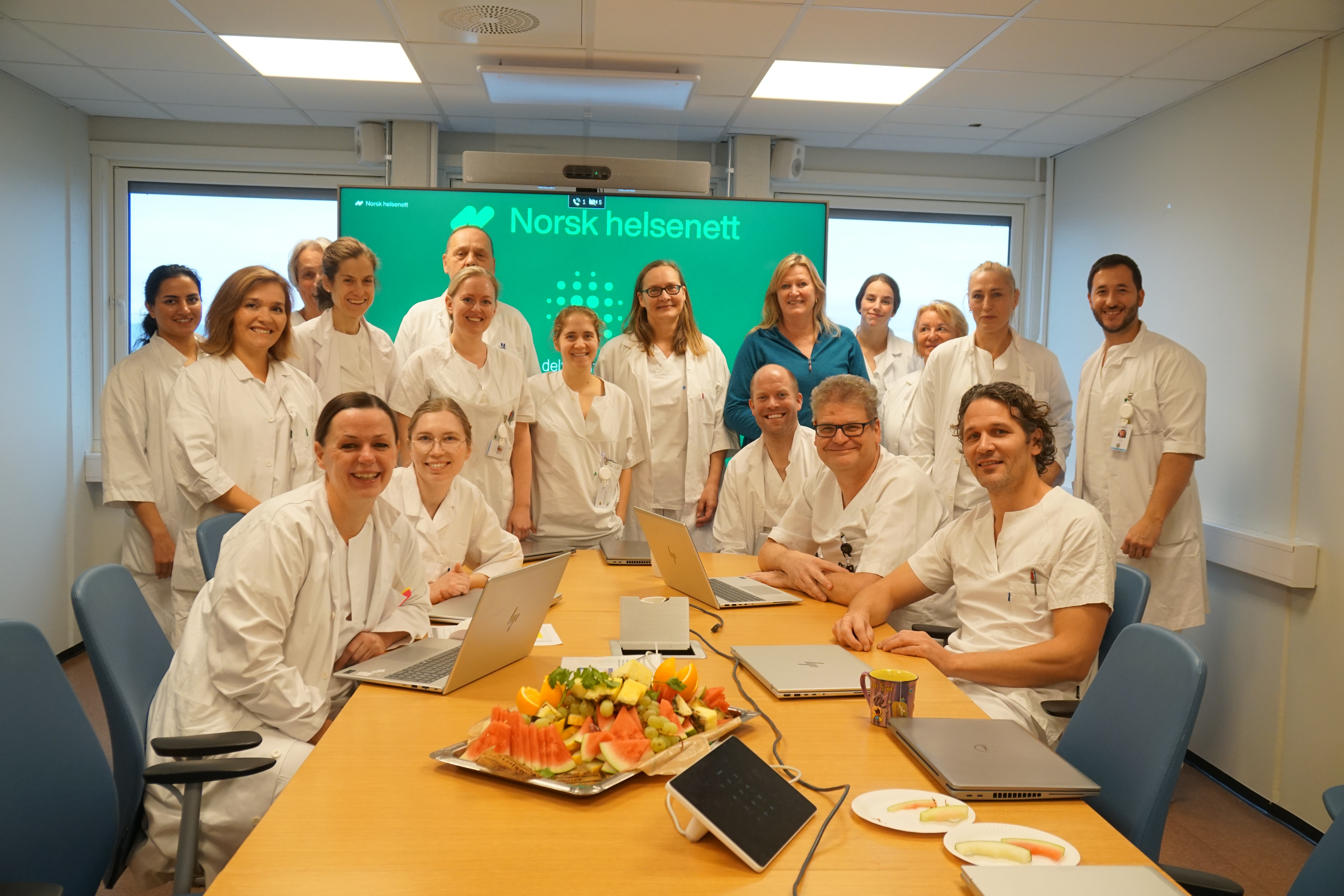 Gruppebilde med leger som sitter og står rundt et bord. Alle smiler til kameraet