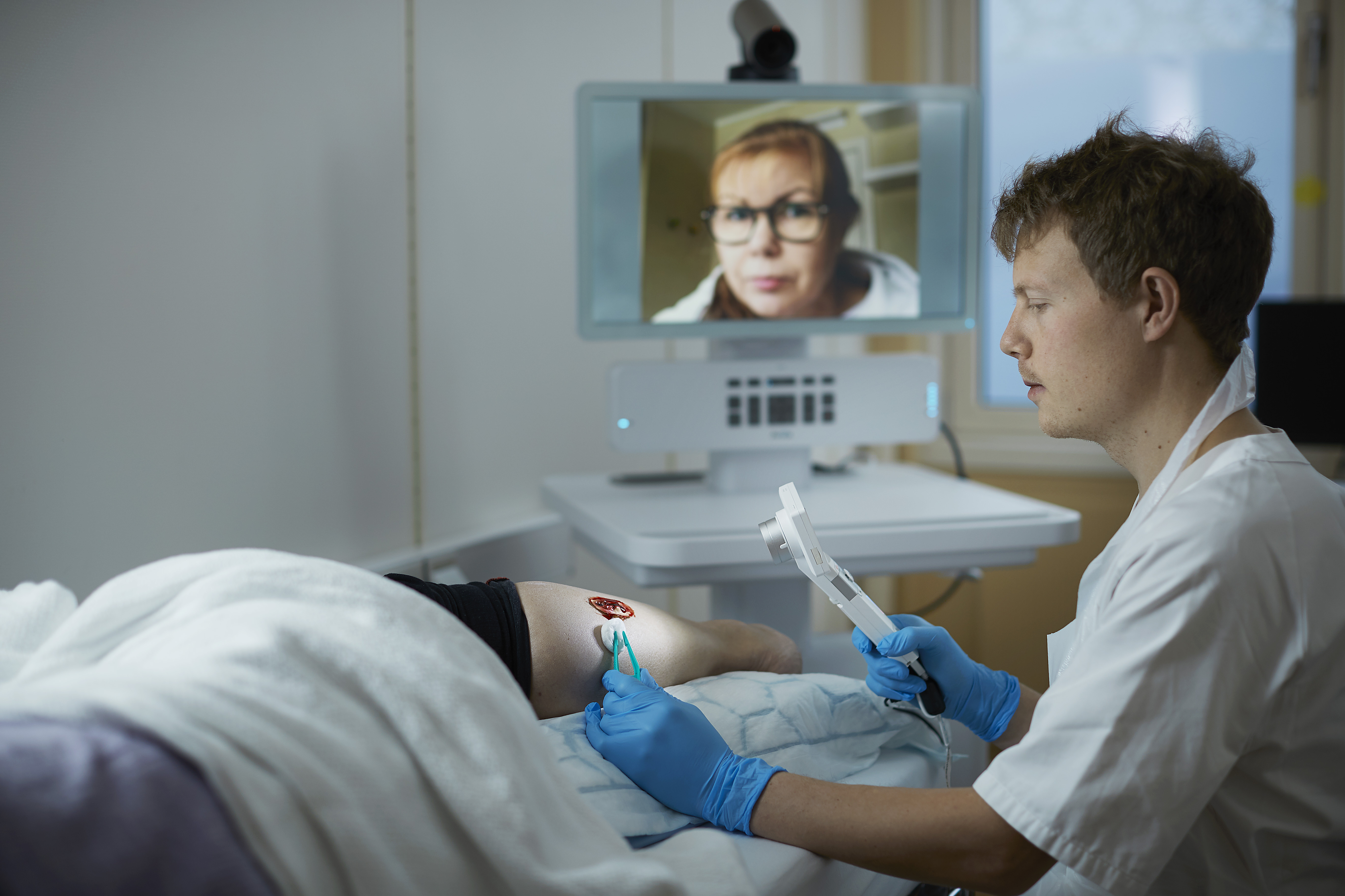 En sykepleier steller et sår, mens han får veiledning av lege over skjerm.