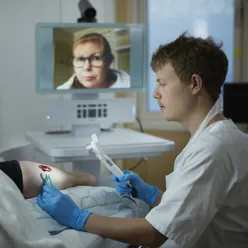 En sykepleier steller et sår, mens han får veiledning av lege over skjerm.