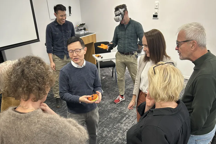 En gruppe forskere står i en ring mens en professor viser en digital sensor. Bak dem står to forskere som tester en hanske og VR-briller